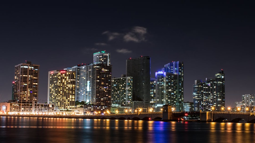Miami skyline at night with illuminated skyscrapers and bridge reflecting on the water.