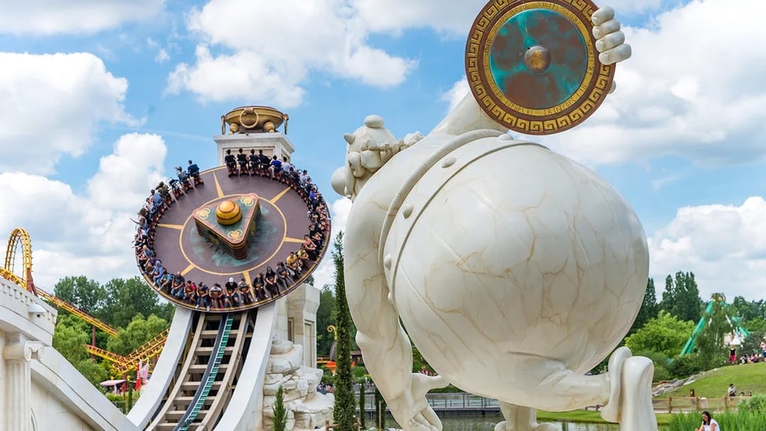Visitors on a spinning ride at Parc Astérix, Paris, with a large statue in the foreground.