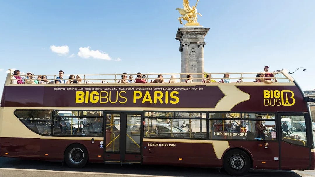 Open-top Big Bus Paris tour passing Pont Alexandre III with tourists on board.
