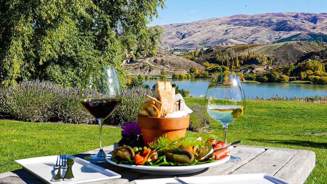 Gourmet platter and wine glasses on a table overlooking a scenic lake near Queenstown.