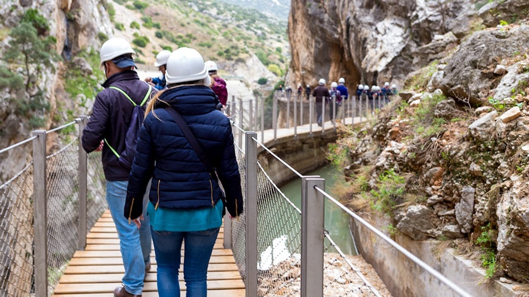 Visitors walking on Caminito del Rey path with helmets, surrounded by cliffs and river in Spain.