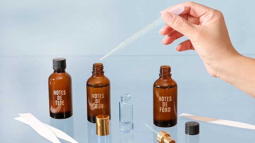 Hand using dropper with perfume bottles labeled "Notes de Tête," "Notes de Cœur," "Notes de Fond" in Grasse workshop.