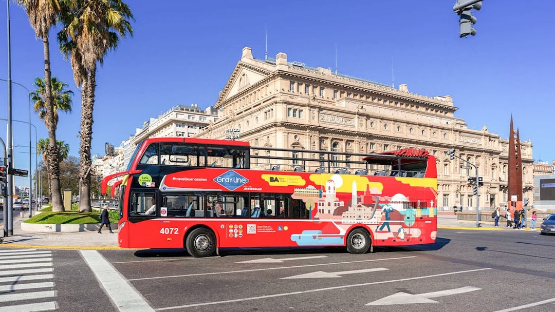 Red double-decker bus in front of Teatro Colón, Buenos Aires, on a sunny day.