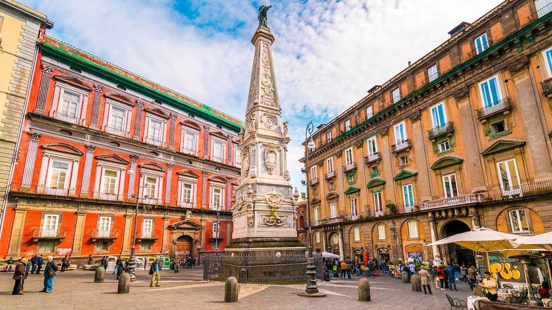 Tourists in Naples exploring historic square with ornate obelisk and colorful buildings.