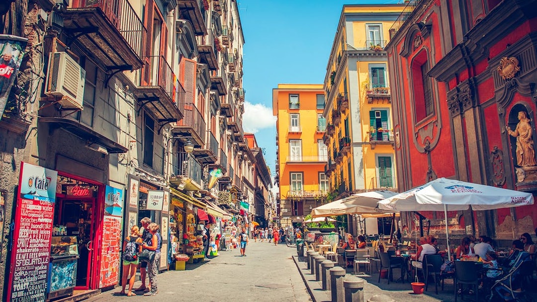 Tourists walking through a vibrant street in Naples with colorful buildings and outdoor cafes.