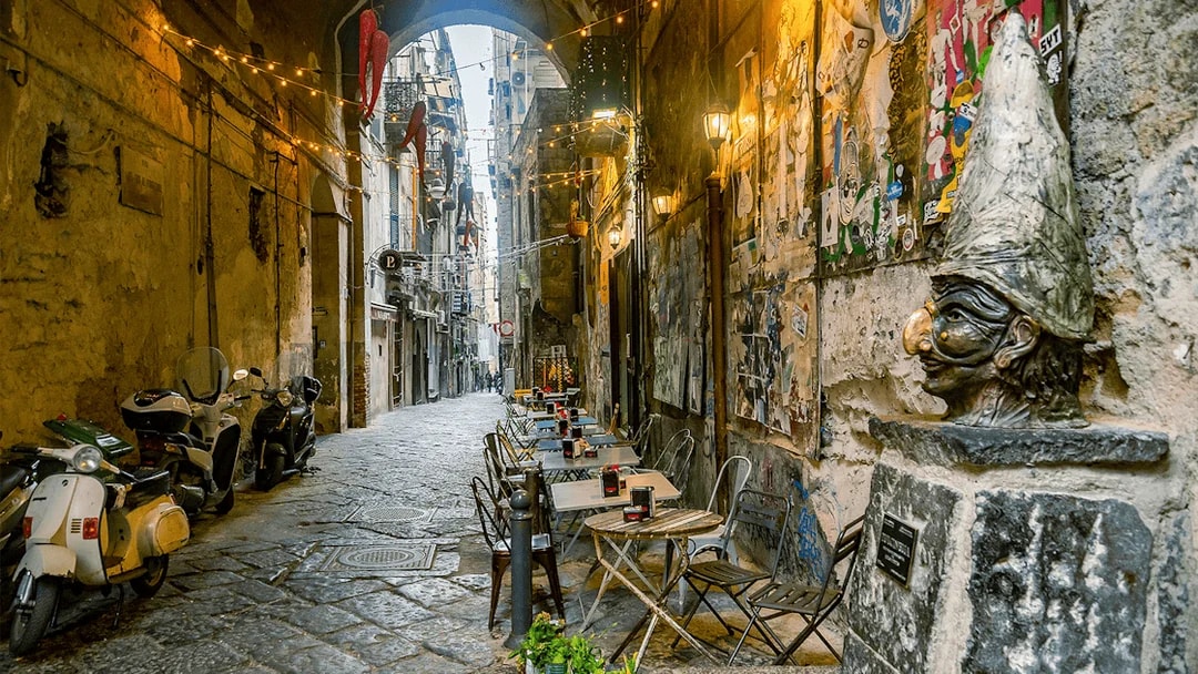 Narrow street in Naples with outdoor café tables and a Pulcinella statue.