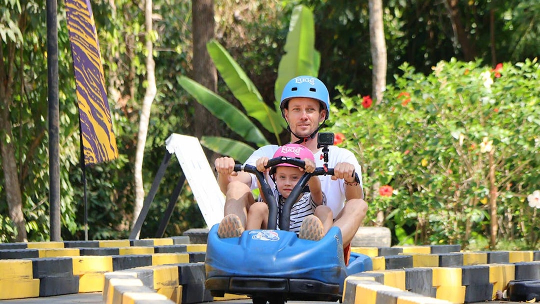 Father and child riding a blue go-kart at Hanuman World, surrounded by lush greenery.