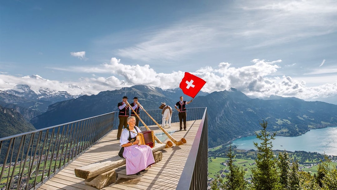 Swiss folk musicians performing at Herder Kulm with alphorns and Swiss flag, overlooking mountains.