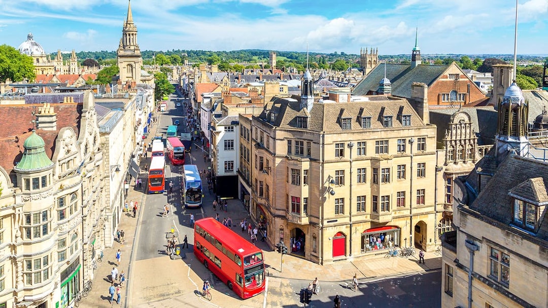 Buses on a busy street in Oxford, with historic buildings and spires in the background.