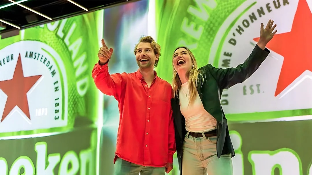 Tourists smiling and pointing at Heineken Experience in Amsterdam.