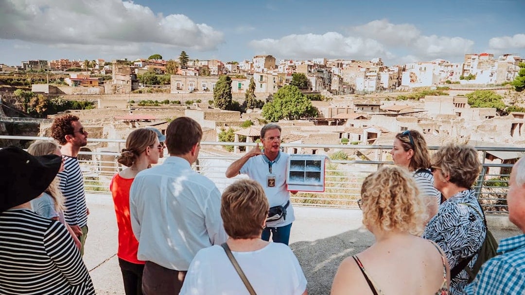 Group listening to a guide at Herculaneum with city ruins in the background.