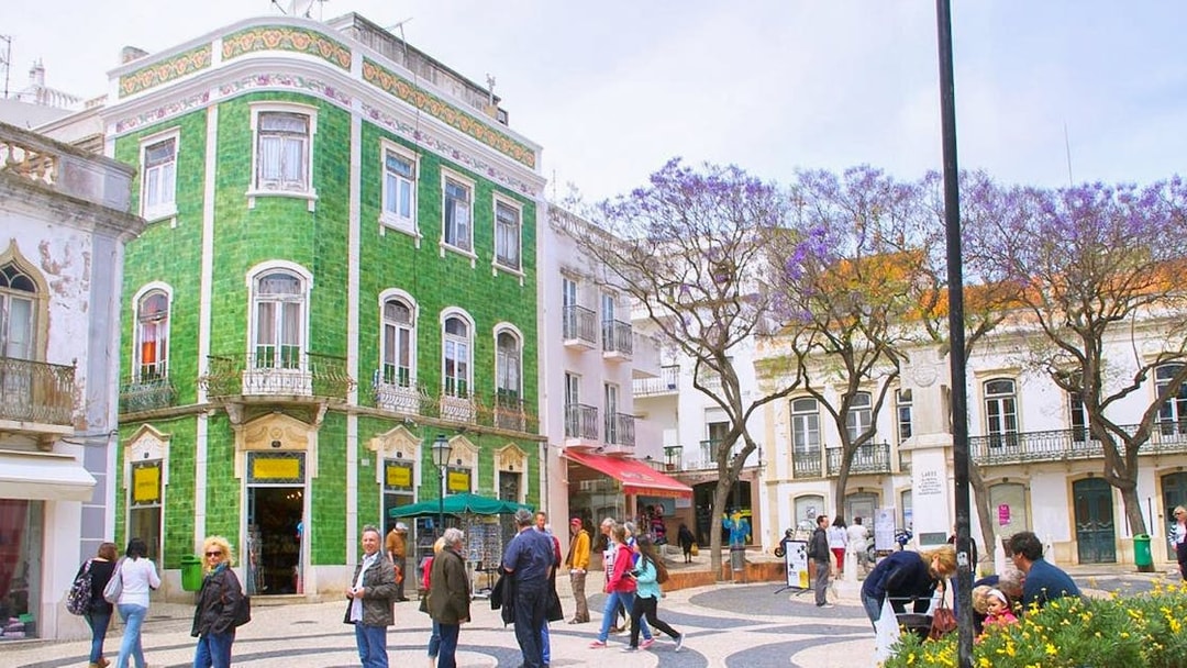 People walking in a historic square with a green-tiled building in Western Algarve.