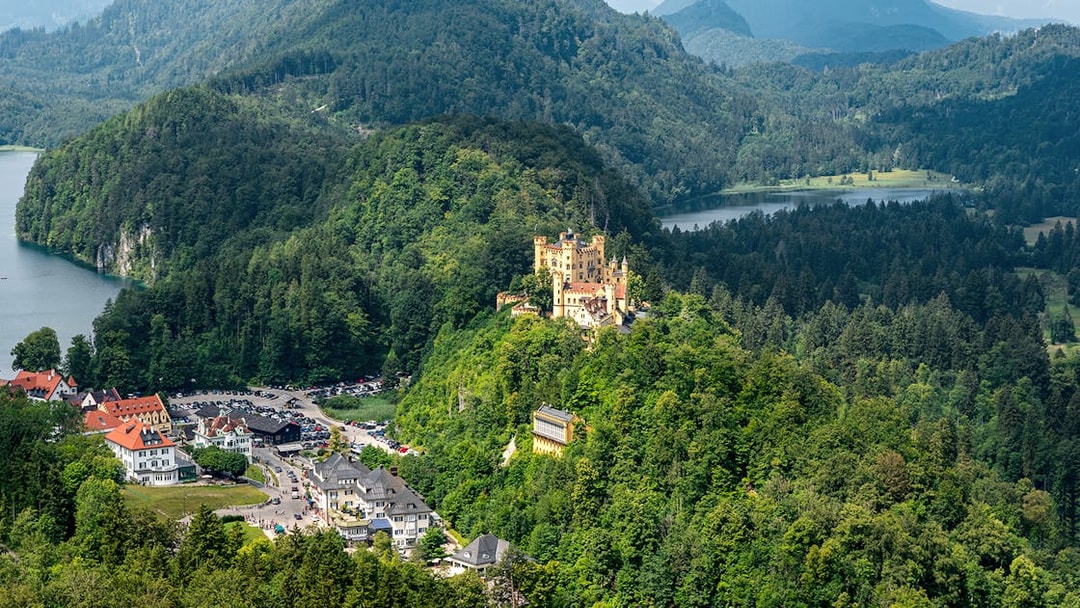 Hohenschwangau Castle surrounded by lush forest and mountains, view of nearby village and lake.
