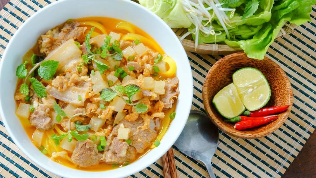 Mi quang noodle dish with pork, herbs, and lime on a woven mat.