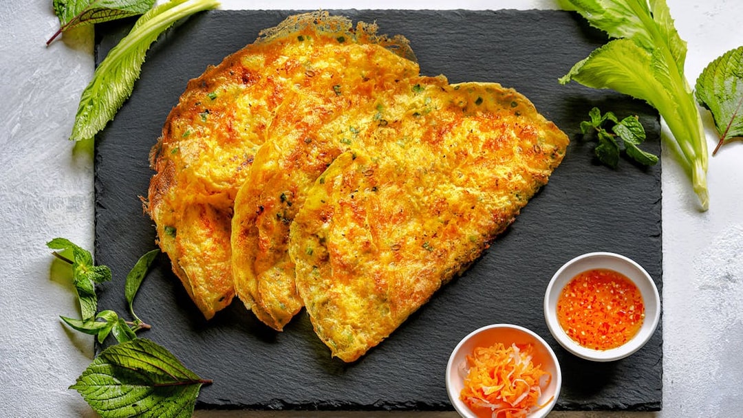 Banh Xeo Vietnamese crepe pancake with herbs and dipping sauce on slate plate.