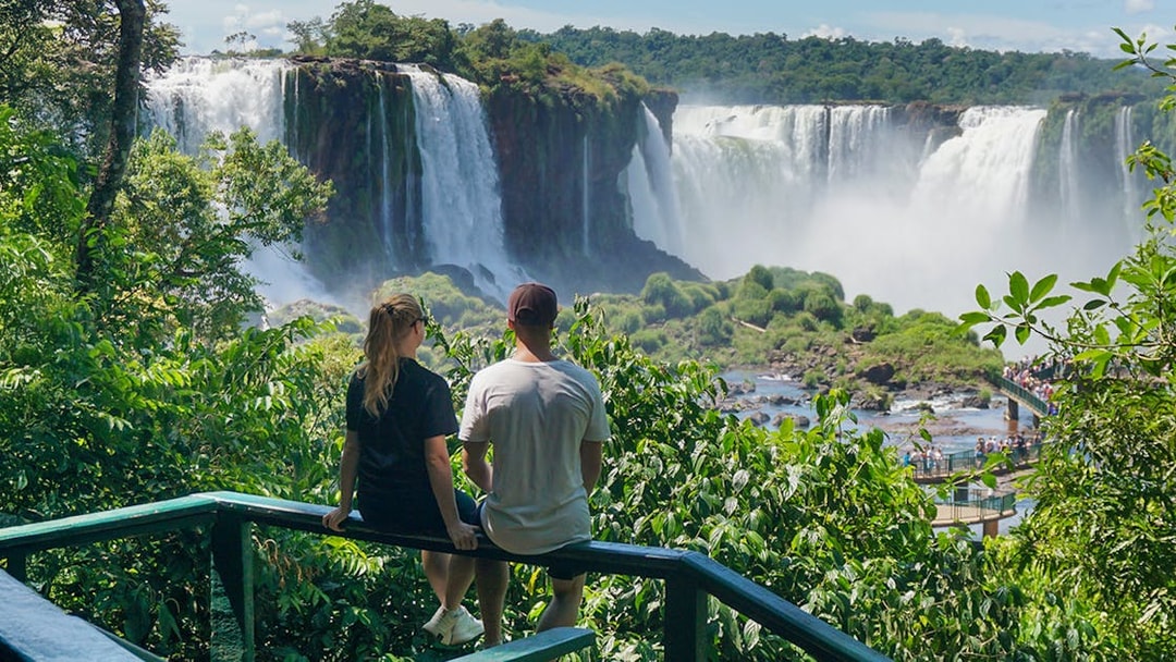 Man and woman viewing Iguazu Falls from a lush green lookout point.