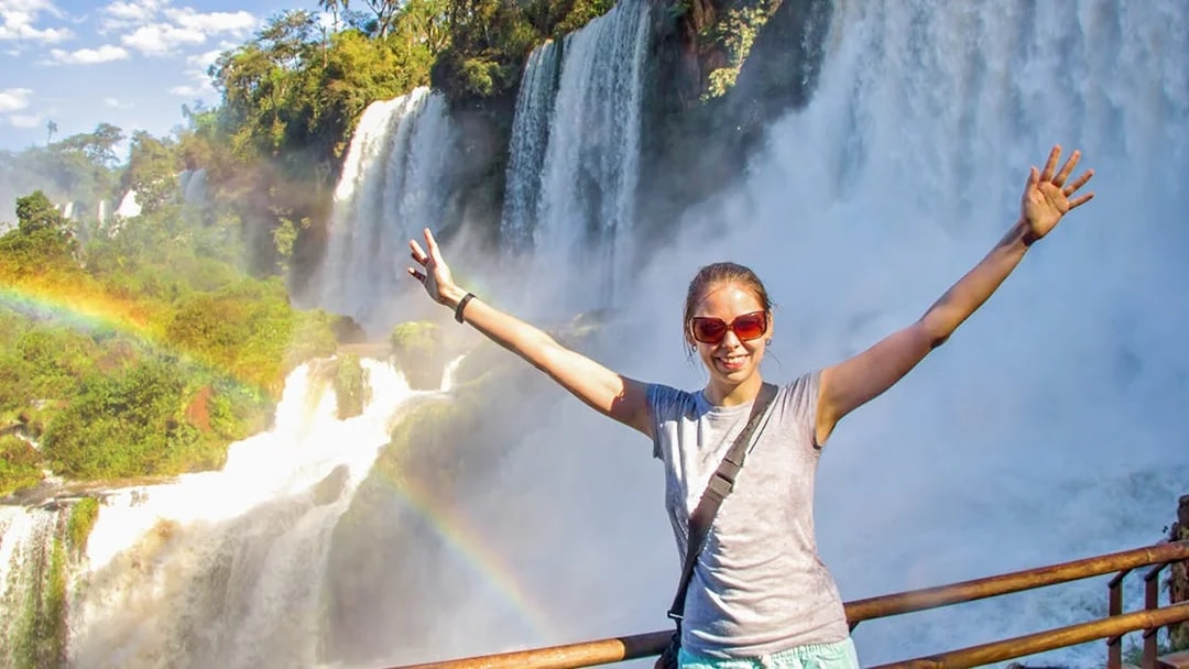 Tourist at Iguazu Falls with arms raised, rainbow visible.