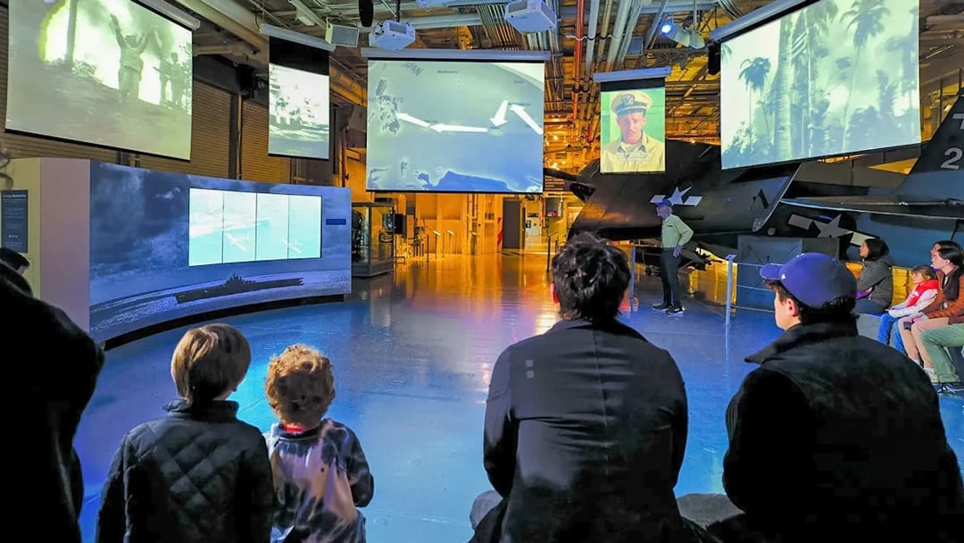 Visitors watching historical footage at a museum exhibit with multiple screens.