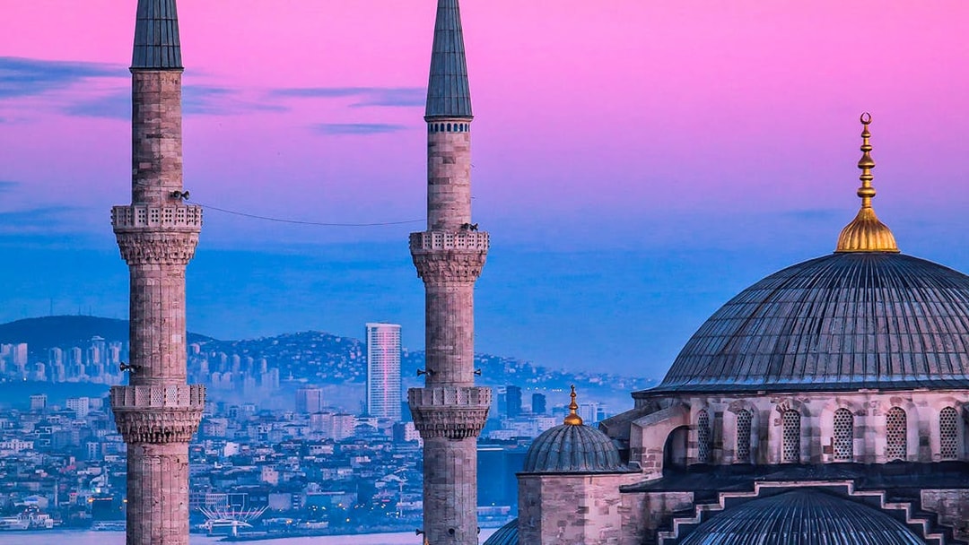 Minarets and dome of a mosque in Istanbul at sunset, with cityscape in the background.
