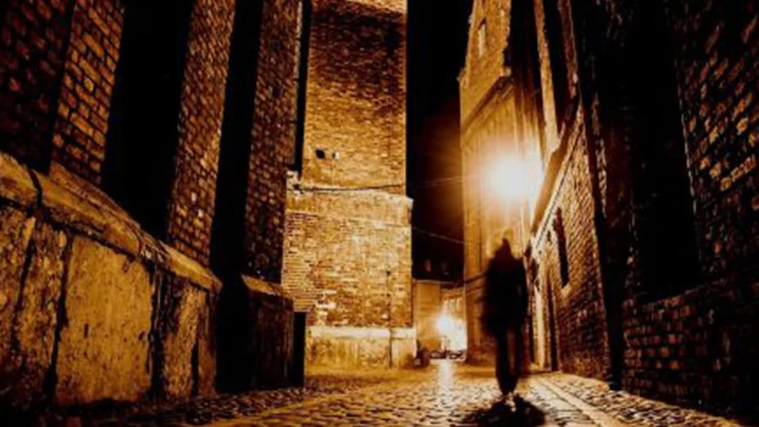 Shadowy figure walking down a dimly lit cobblestone alley in London.