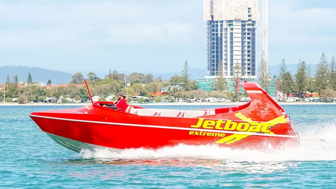 Red jet boat speeding on water near cityscape, part of Ultimate Jet Boat Ride and Surf Lesson.