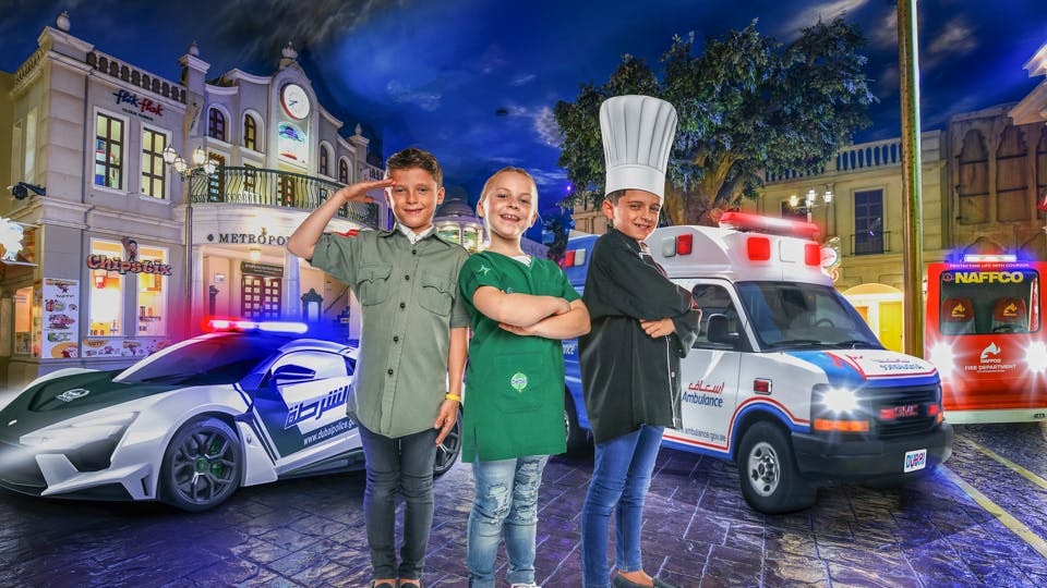 Children in role-play costumes at KidZania Dubai with police car and ambulance in background.