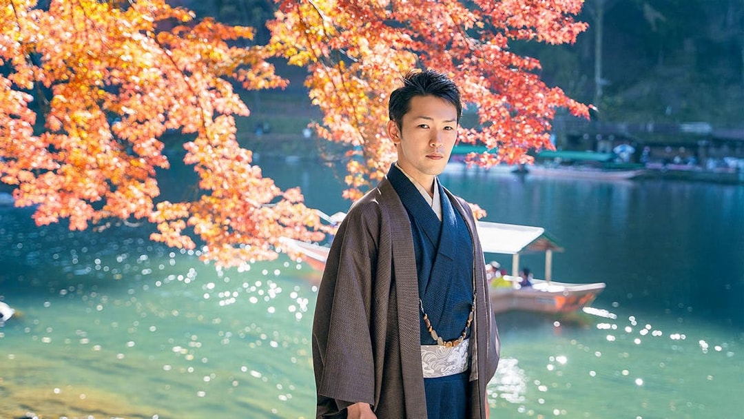 Man in kimono by river with autumn leaves in Kyoto Arashiyama.