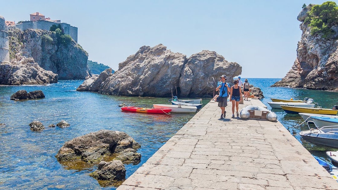 Tourists walking along a stone pier in Dubrovnik with boats and rocky cliffs nearby.
