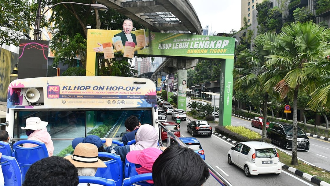 Open-top bus tour in Kuala Lumpur with passengers viewing city streets and greenery.