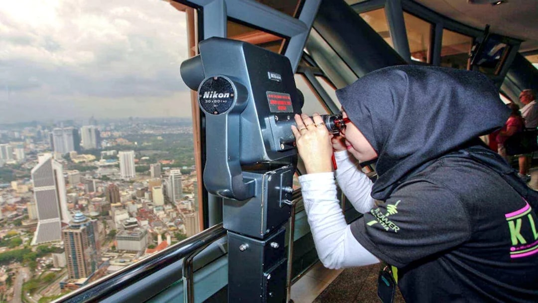 Visitor using binoculars at KL Tower observation deck, overlooking Kuala Lumpur cityscape.