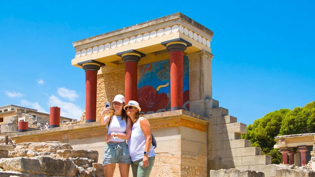 Visitors taking a selfie at Knossos Palace, Crete, with ancient fresco and columns in view.