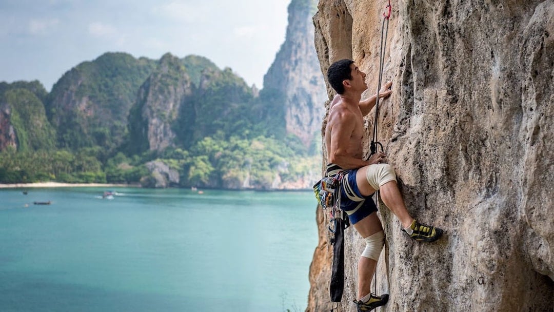 Rock climber scaling limestone cliff at Railay Beach, Krabi, with ocean and cliffs in background.