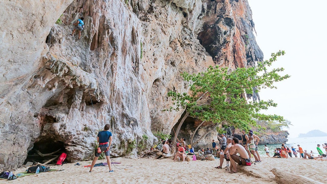 Rock climbers at Railay Beach, Krabi, scaling limestone cliffs with onlookers nearby.