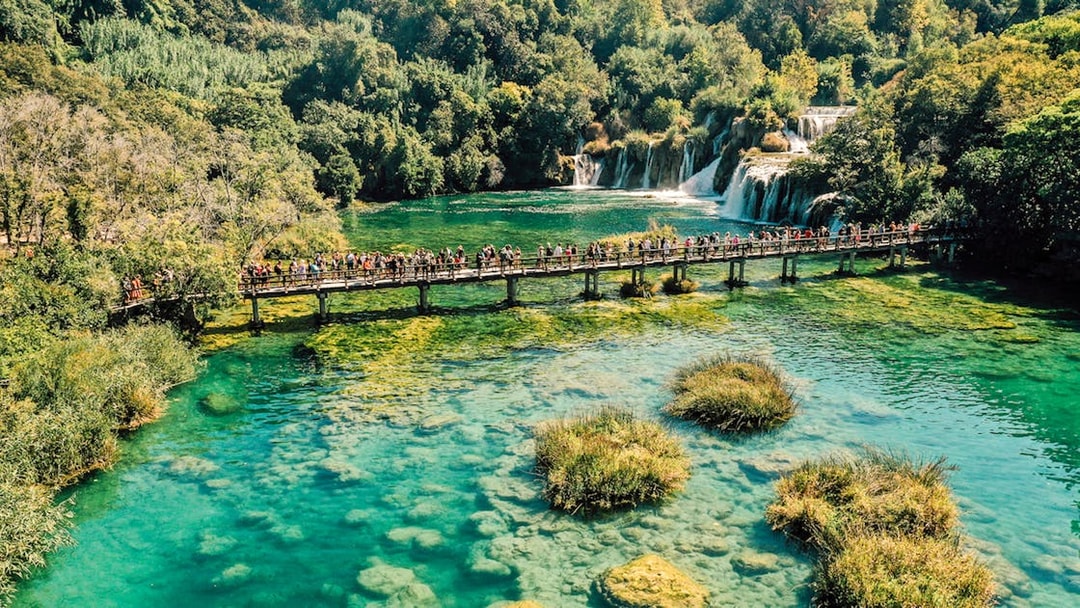 Visitors on a bridge over turquoise water at Krka National Park, Croatia, with waterfalls in the background.