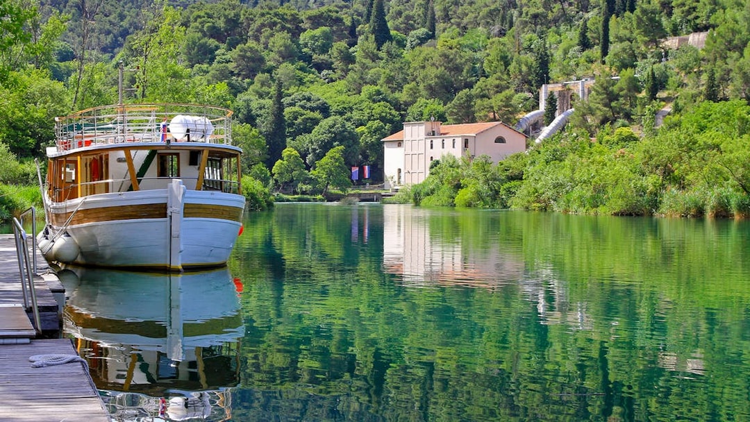 Boat docked on Krka National Park river with lush greenery and historic building in background.