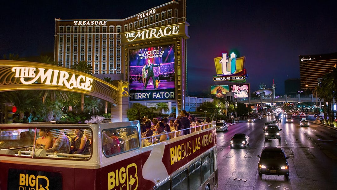 Open-top bus tour passing The Mirage and Treasure Island in Las Vegas at night.