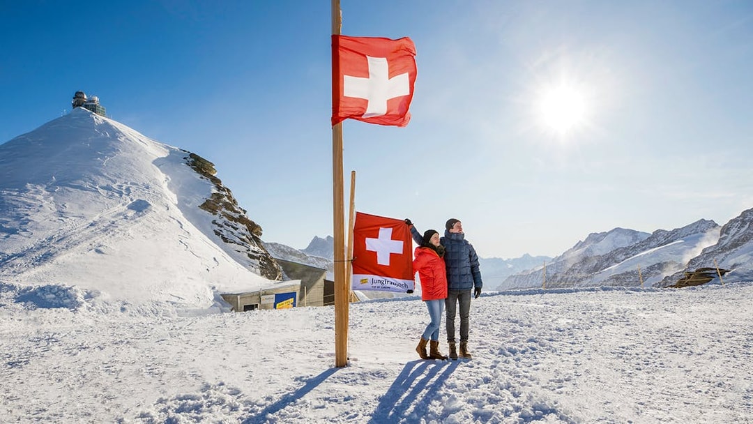 Visitors at Jungfraujoch Plateau with Swiss flags and snowy mountain backdrop.