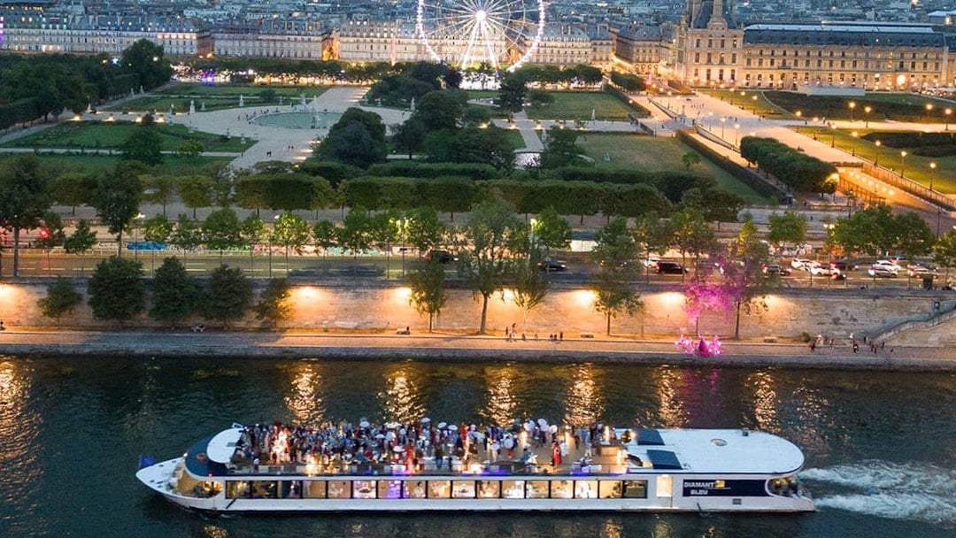 Seine River dinner cruise boat with guests, Paris Ferris wheel and city lights in background.