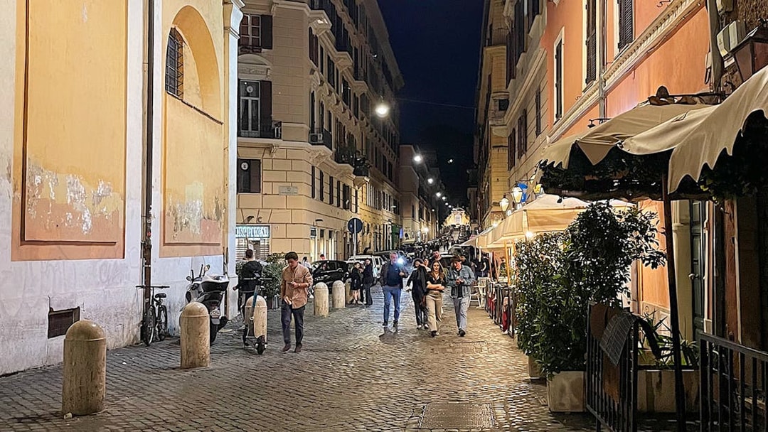 Cobblestone street in Rome at night with people walking and outdoor cafes.