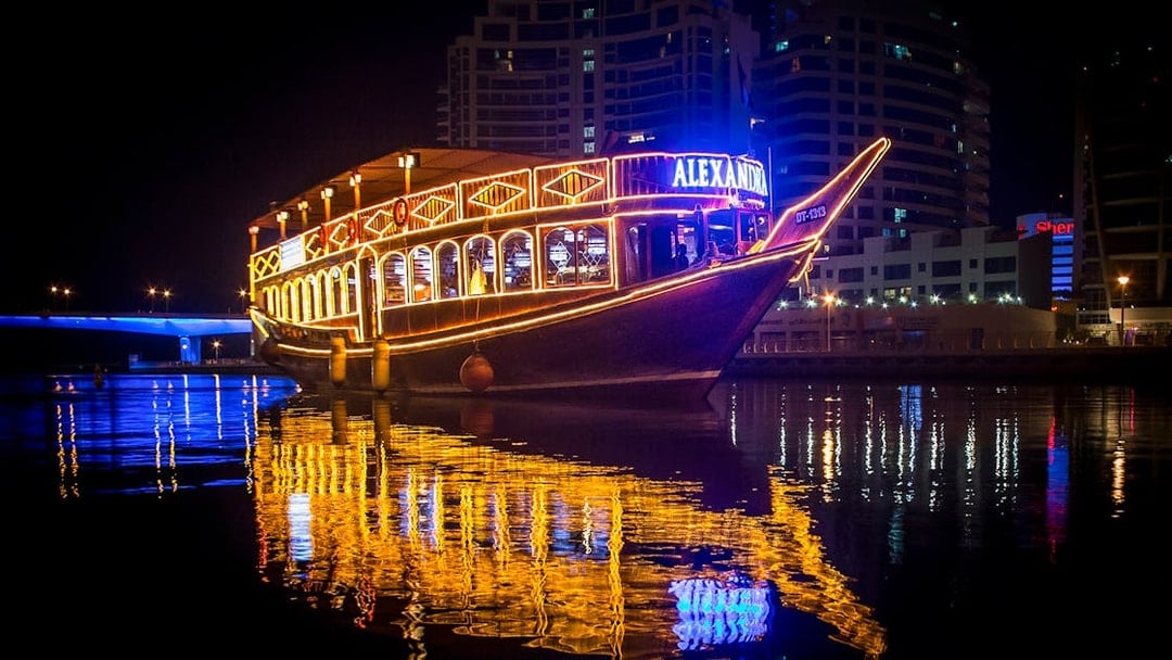 Alexandra Dhow Cruise lit up at night in Dubai Marina, reflecting on the water.