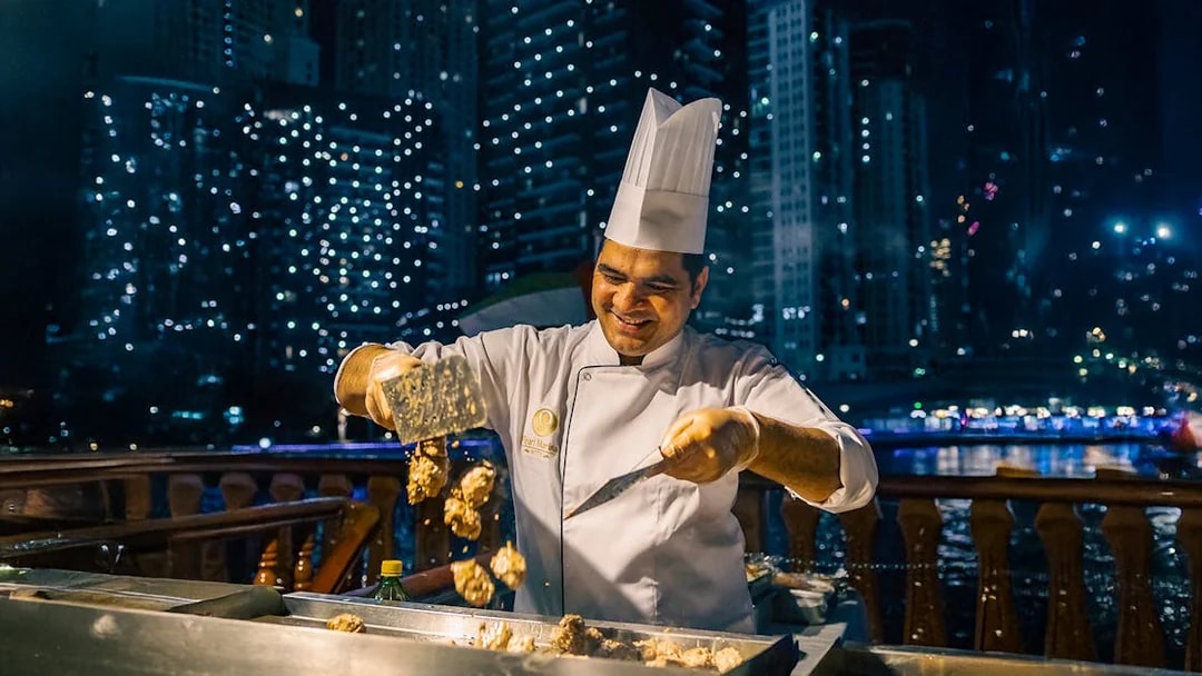 Chef preparing food on a Dubai Marina dhow cruise with city lights in the background.