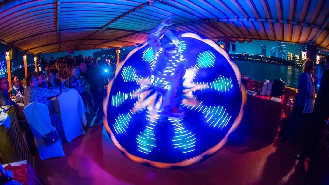 Tanoura dancer performing on Alexandra VIP Dhow Cruise with city skyline in background.
