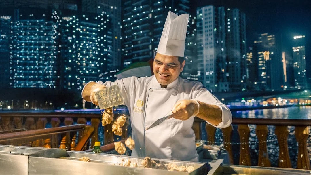 Chef grilling meat on Alexandra VIP Dhow Cruise with city skyline in background.