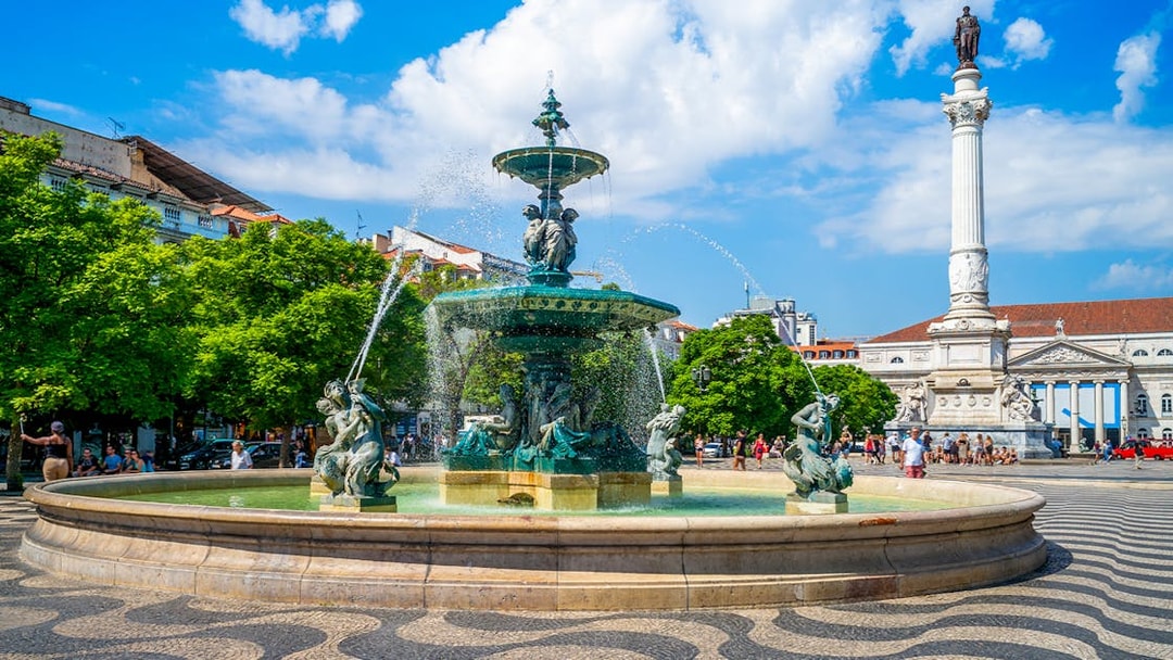 Fountain and column in Rossio Square, Lisbon, with people and trees in the background.