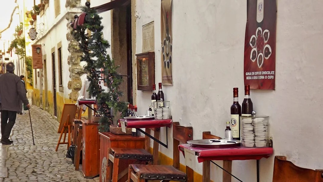 Cobblestone street in Óbidos with outdoor café tables and ginjinha bottles.