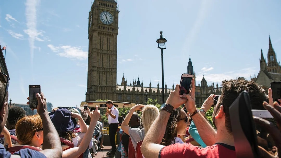 Tourists on a Big Bus near Big Ben in London, taking photos of the landmark.
