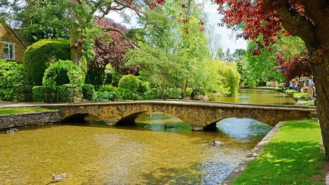 Stone bridge over a tranquil river in Bourton-on-the-Water, England, surrounded by lush greenery.