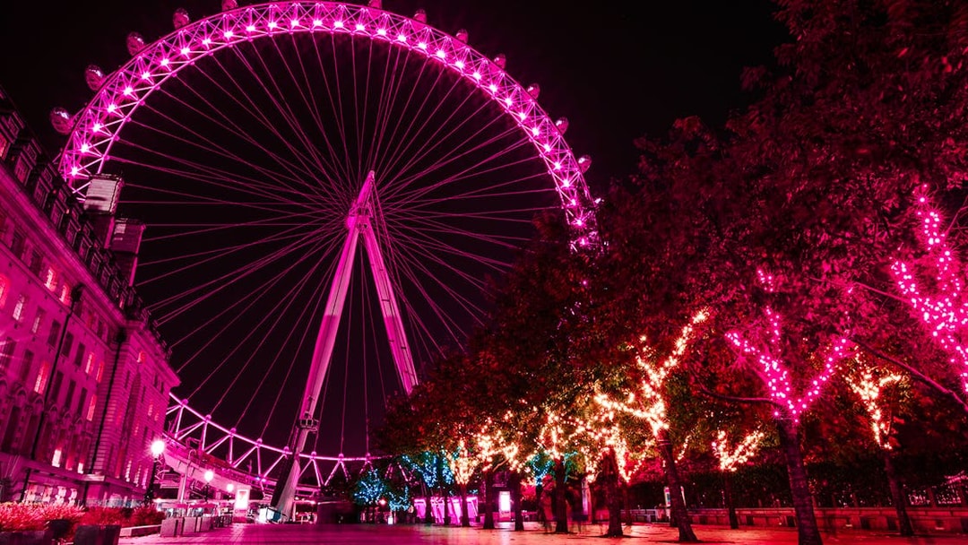 London Eye illuminated at night with vibrant pink lights, trees adorned with festive lights.