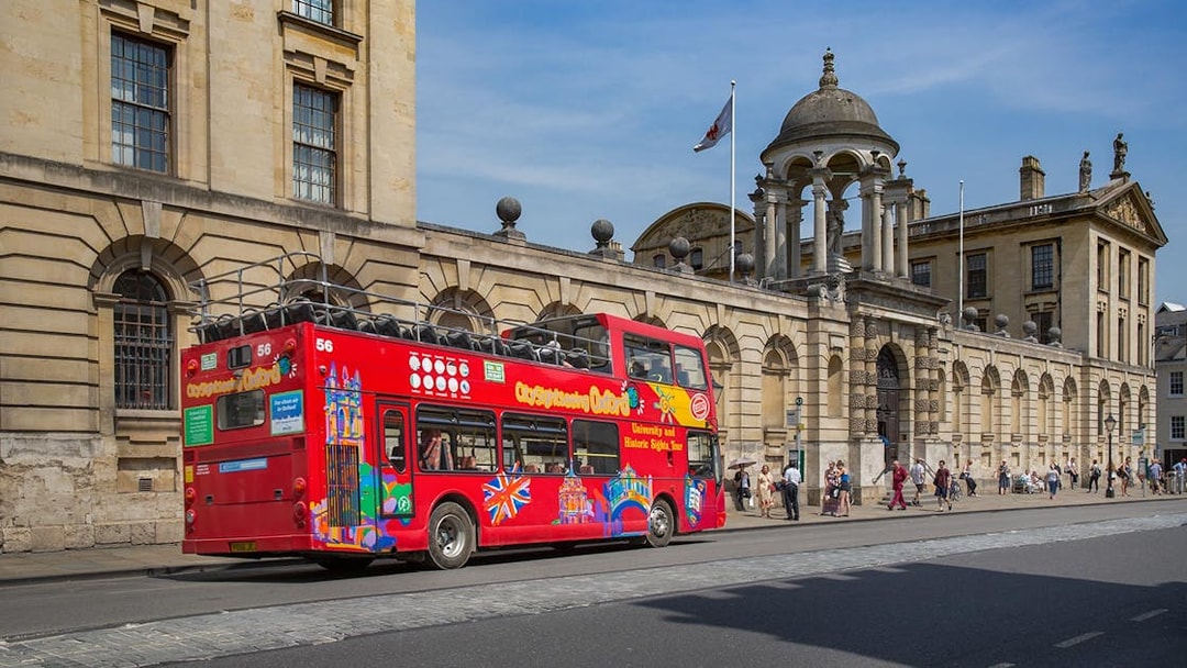 Red double-decker bus on Oxford street near historic building, part of Oxford by Rail day tour from London.