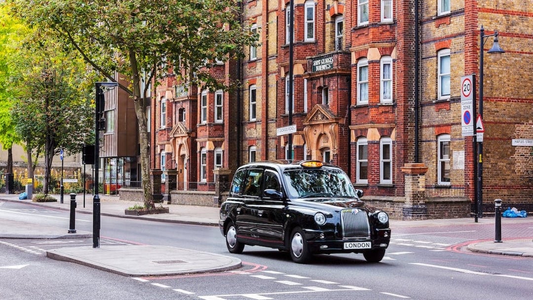 Black taxi driving on a London street with historic buildings in the background.